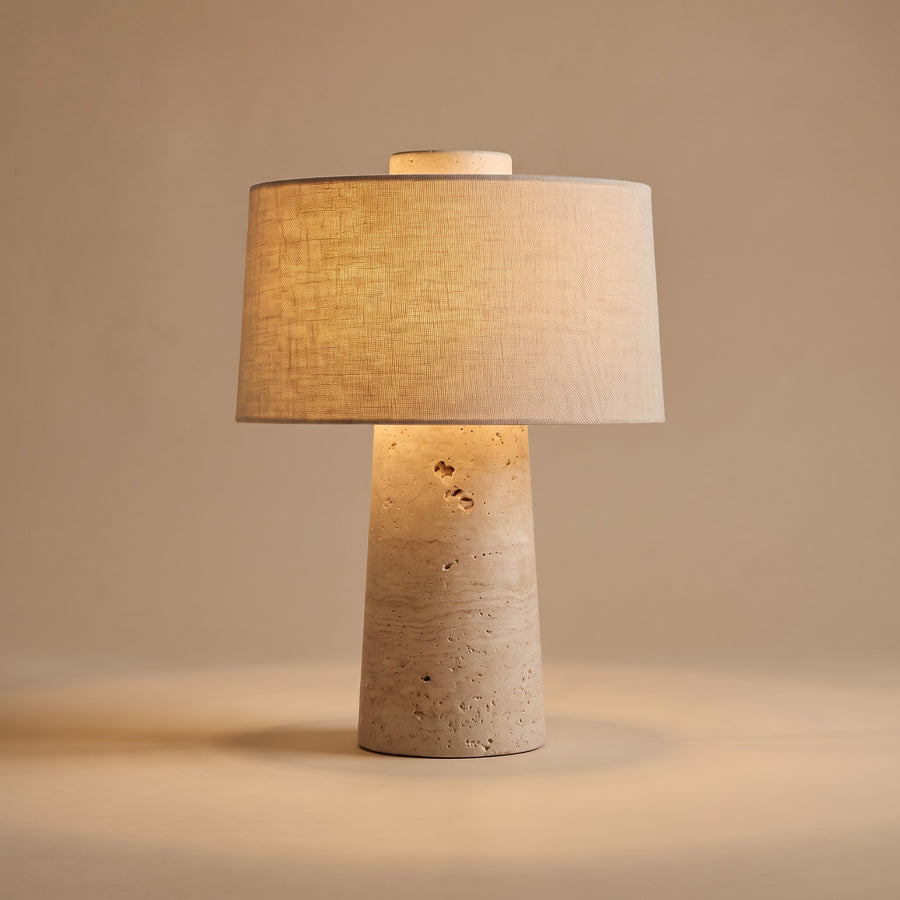 Tellur travertine and fabric table lamp, lit, against a warm beige backdrop