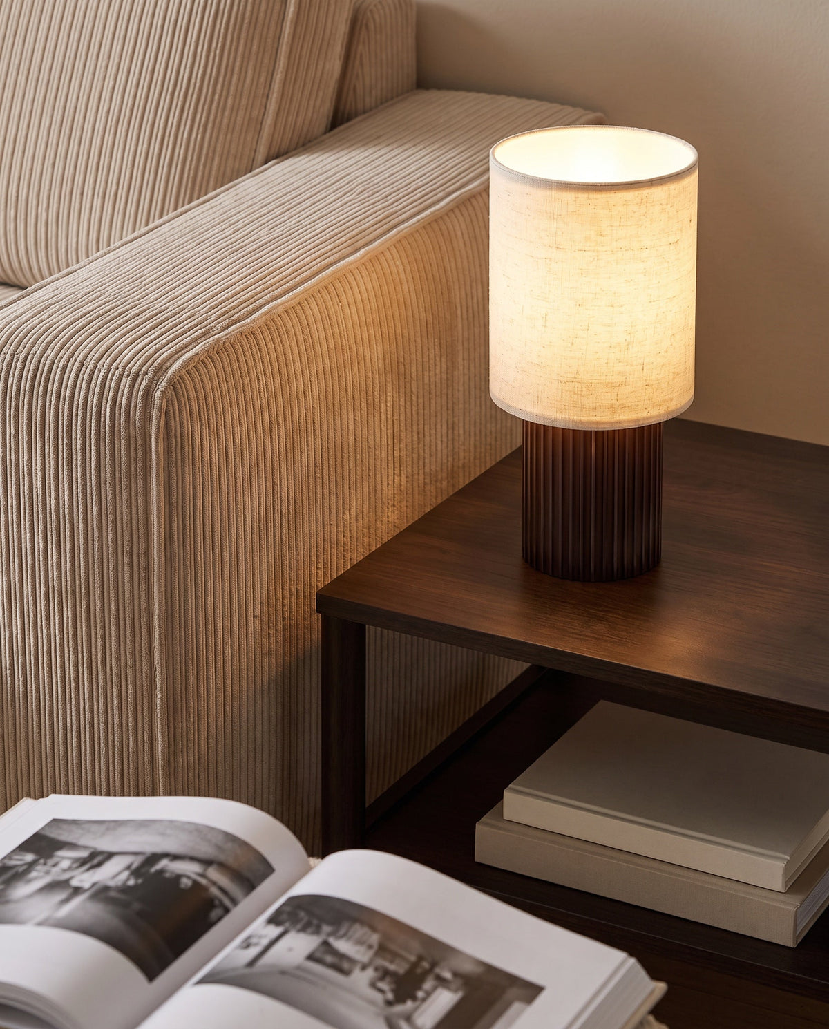 Colonne aluminum and linen table lamp, lit, on a dark wood side table beside a corduroy sofa