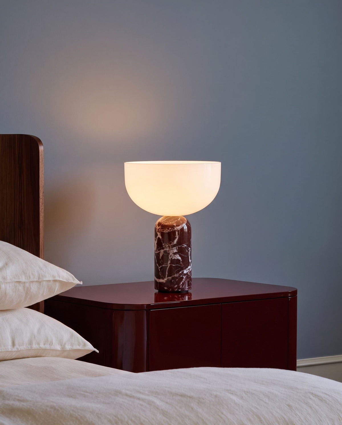 New Works marble and glass table lamp, lit, on a burgundy nightstand beside a linen bed