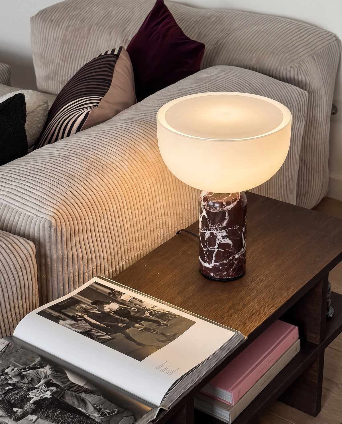 New Works marble and glass table lamp, lit, its opal glass bowl on a red marble base, on a side table beside a corduroy sofa