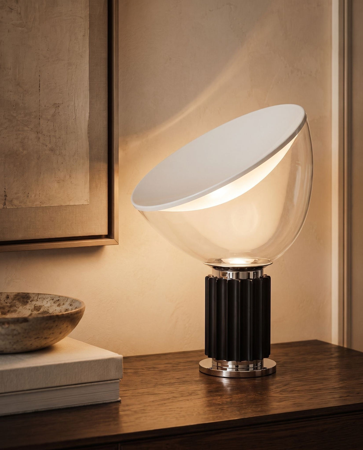 Zenith glass and metal table lamp, lit, its tilted clear glass bowl on a dark wood console beside framed art and books
