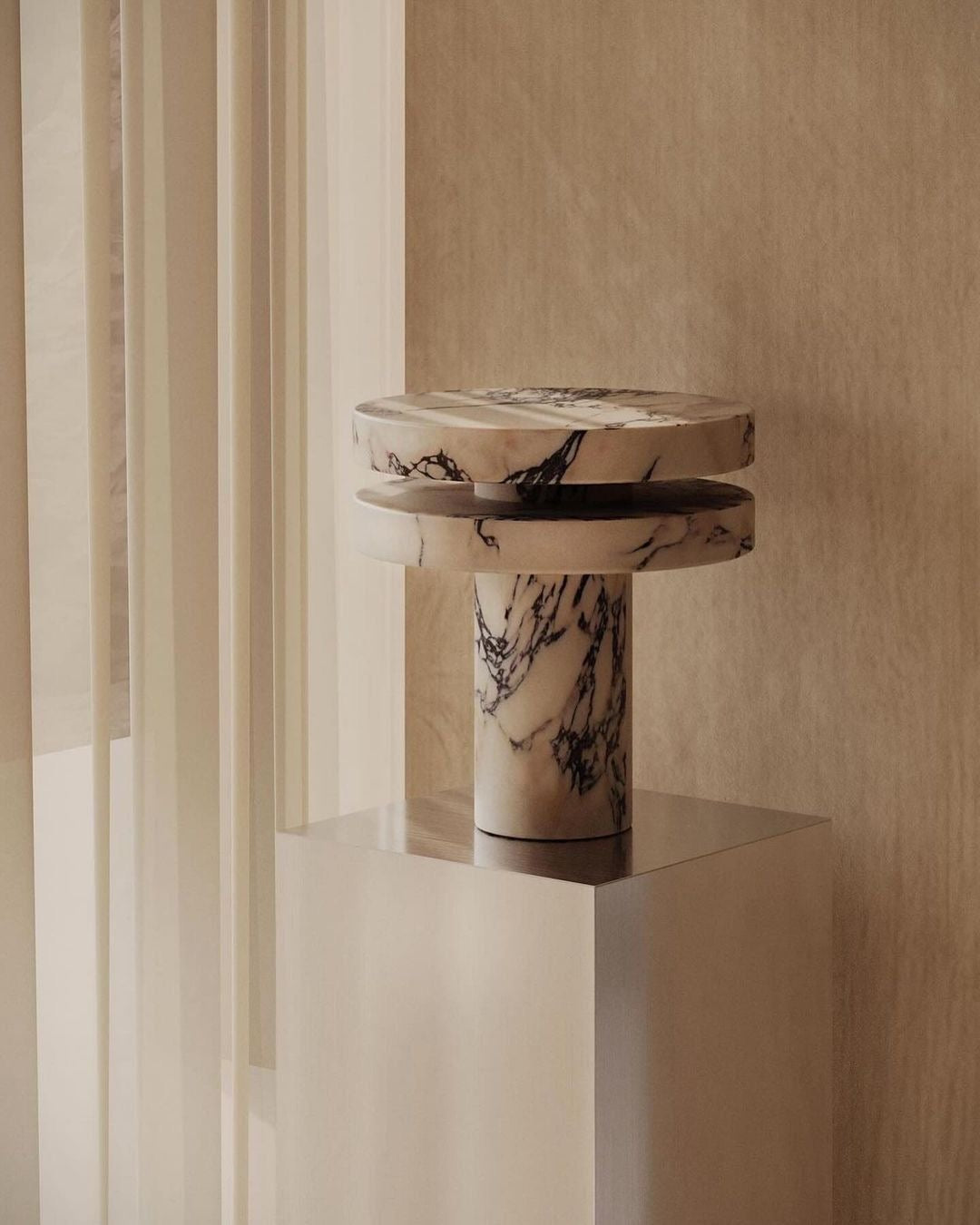 Strates marble table lamp on a white plinth beside sheer curtains, warm beige wall