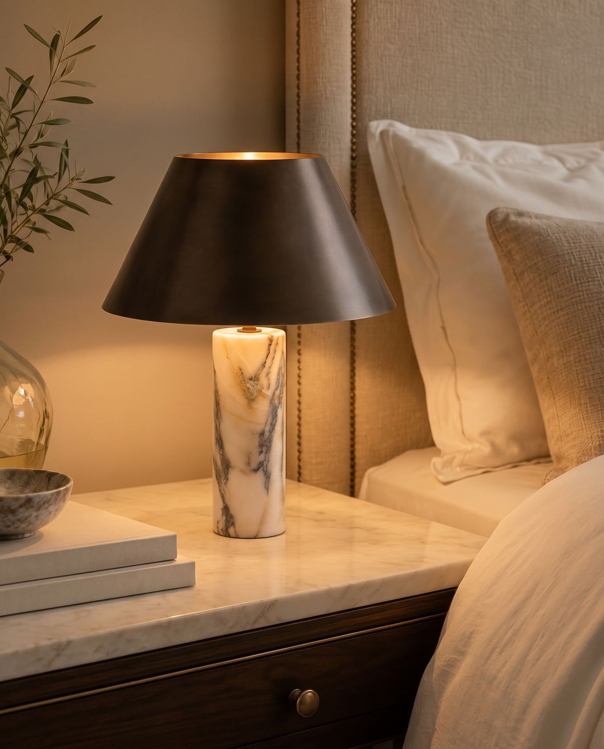 Cline Vesper marble and metal table lamp, lit, beside an upholstered bed