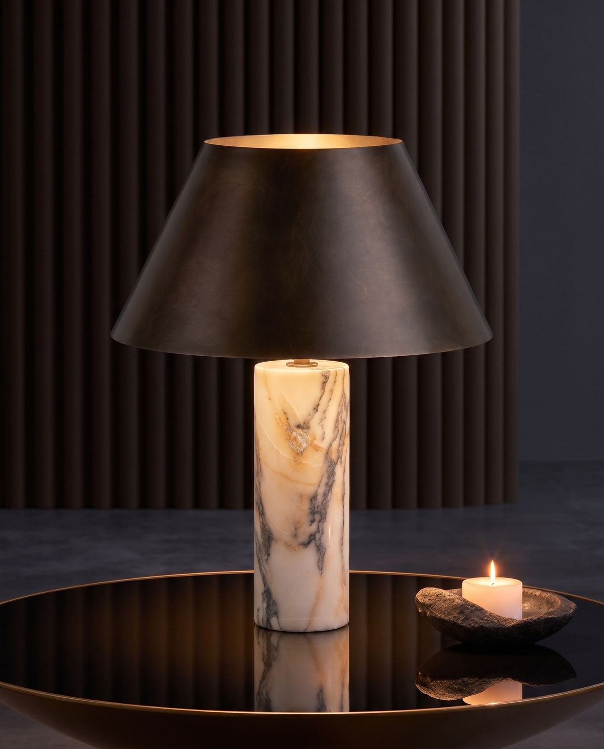 Cline Vesper marble and metal table lamp, lit, on a mirrored round table beside a lit candle