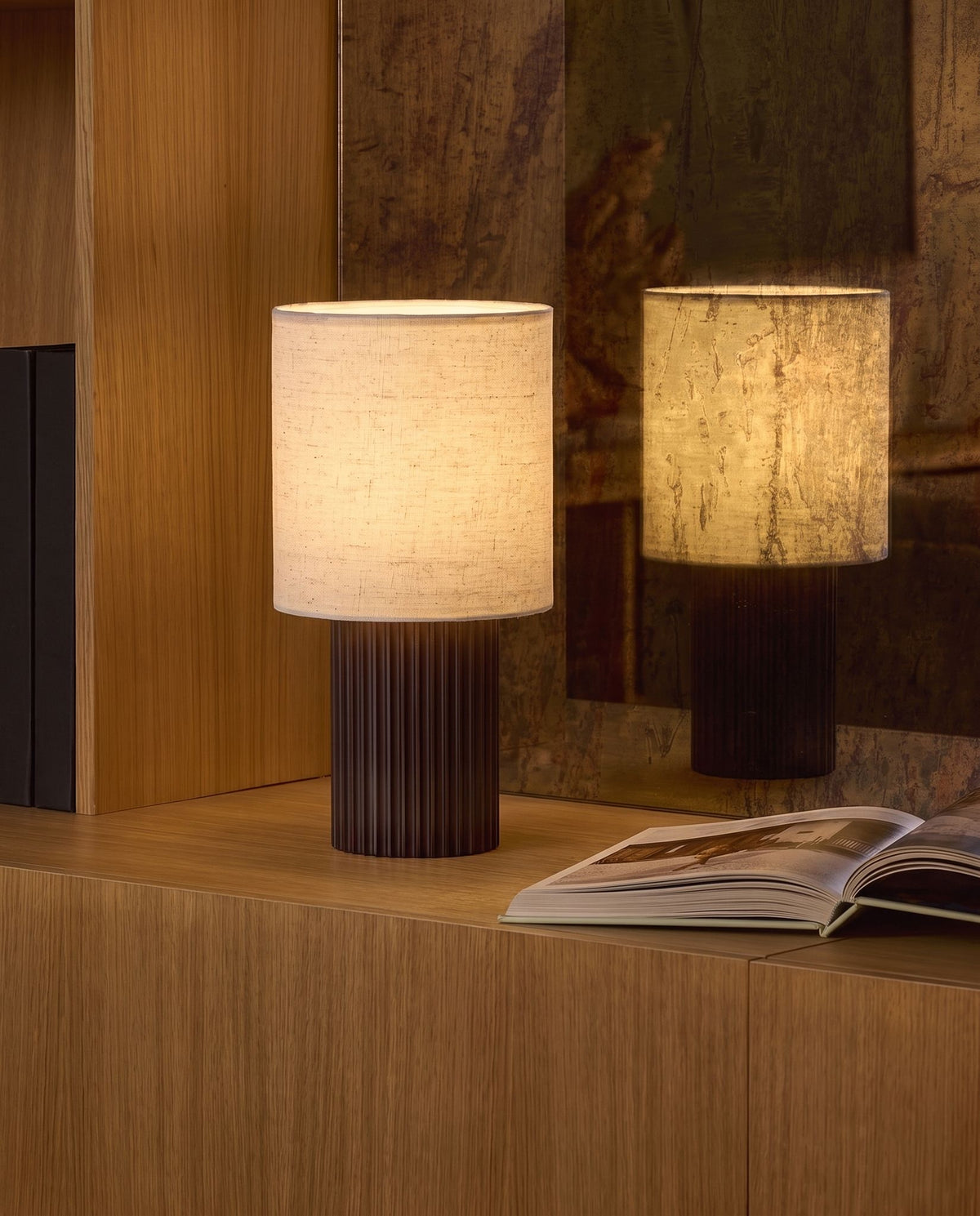 Colonne aluminum and linen table lamp, lit, on oak cabinet, reflected in an antiqued mirror beside open book