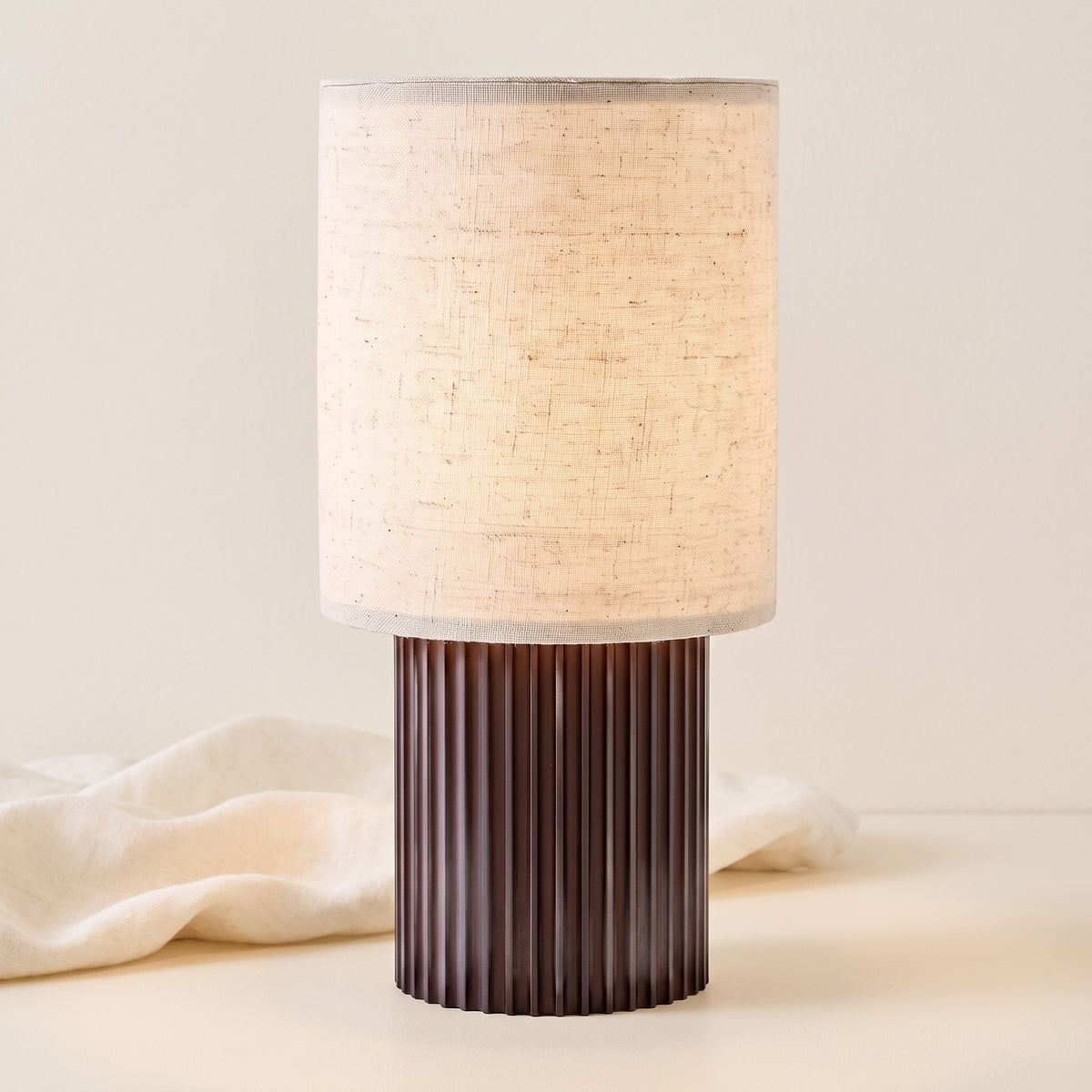 Colonne aluminum and linen table lamp, lit, beside cream fabric on off-white