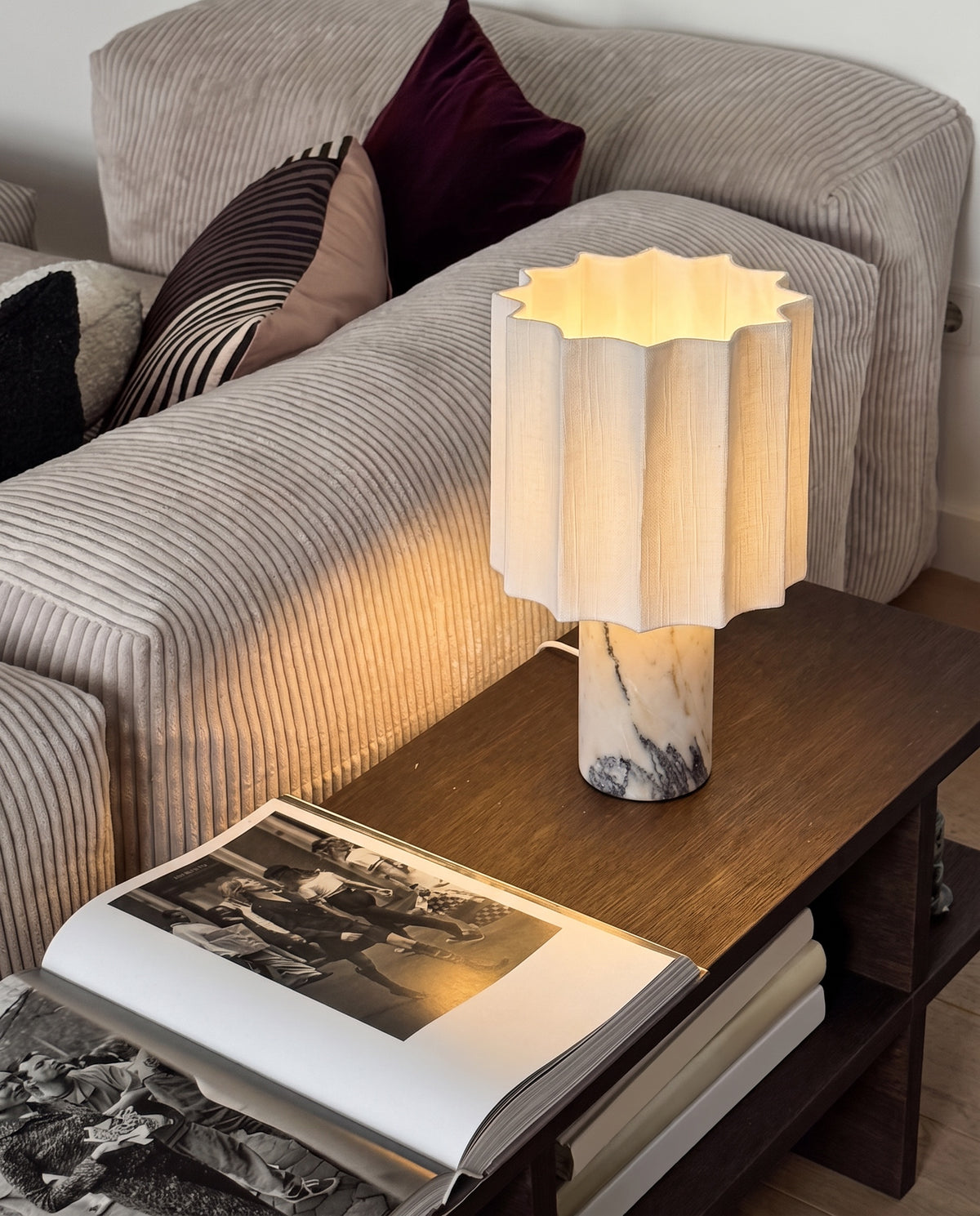 Macara marble and fabric table lamp, lit, on a wood side table next to a corduroy sofa
