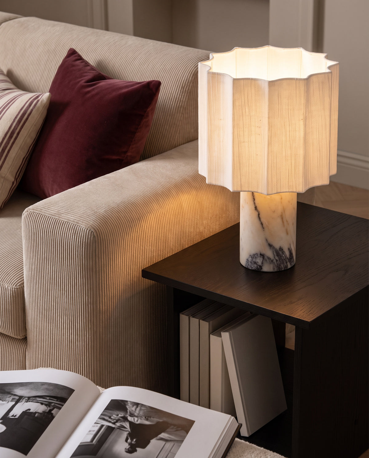 Macara marble and fabric table lamp, lit, on dark side table beside a corduroy sofa with burgundy cushion
