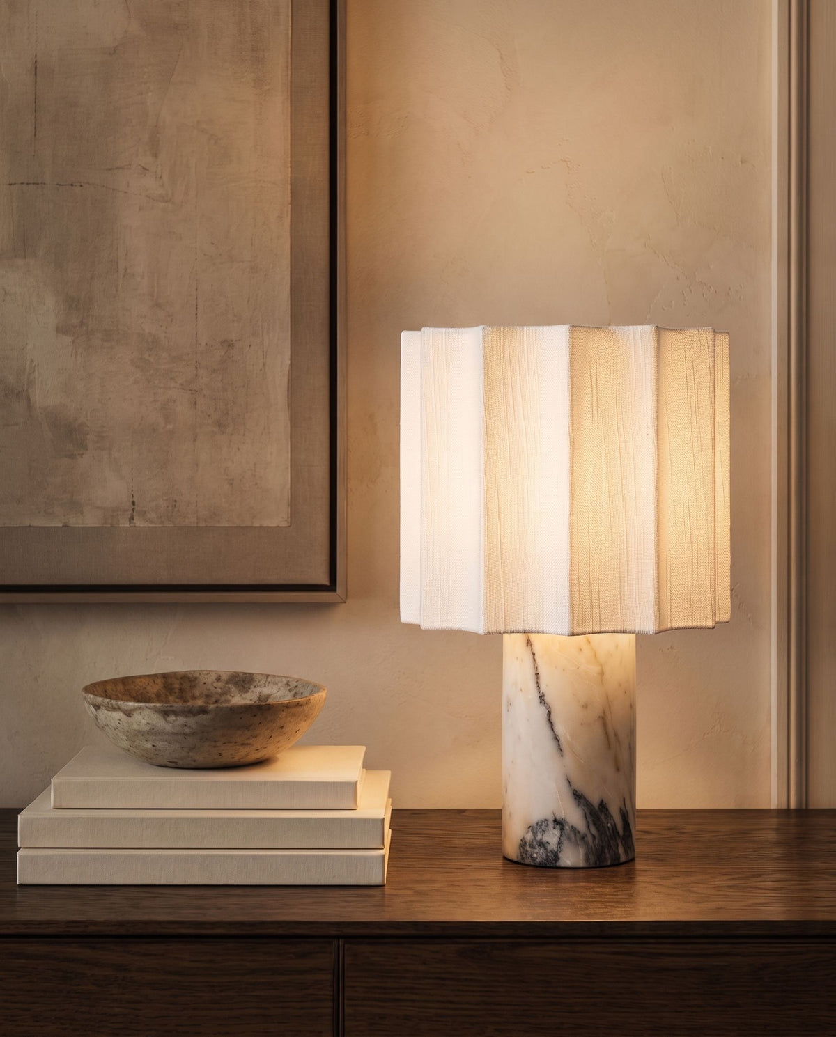 Macara marble and fabric table lamp, lit, on a dark wood dresser beside books and stone bowl