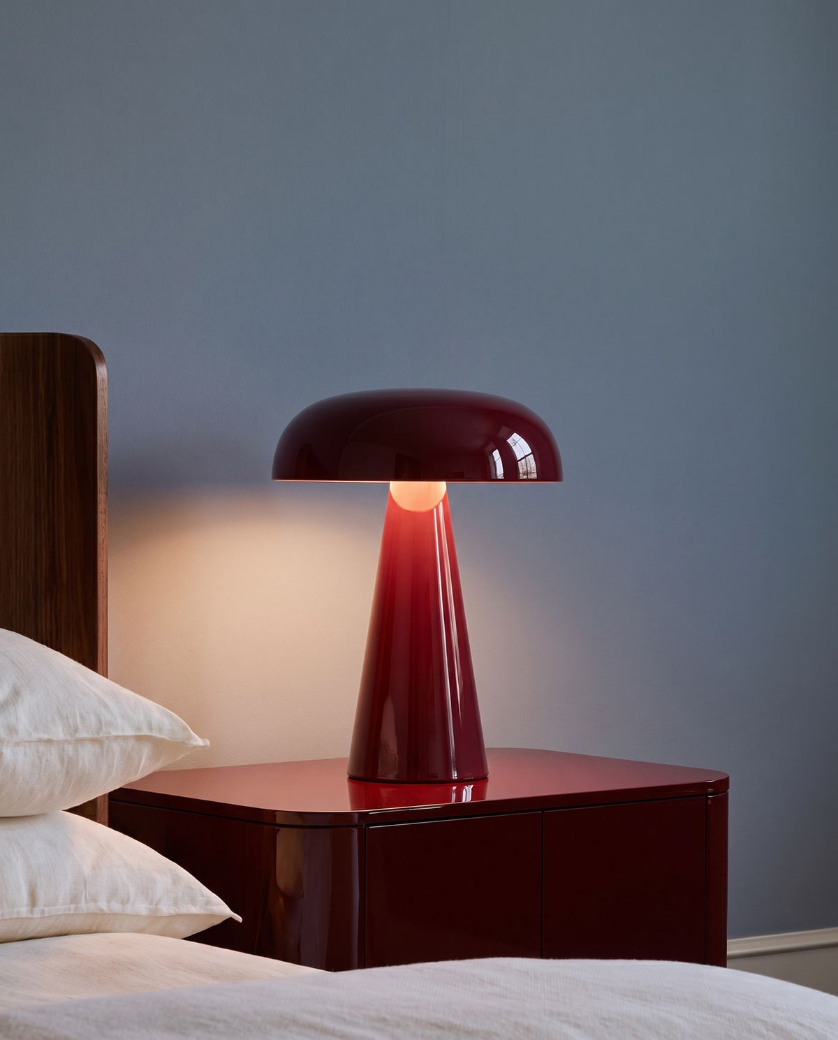 Rubra lacquered table lamp, lit, on a red lacquered nightstand beside a bed