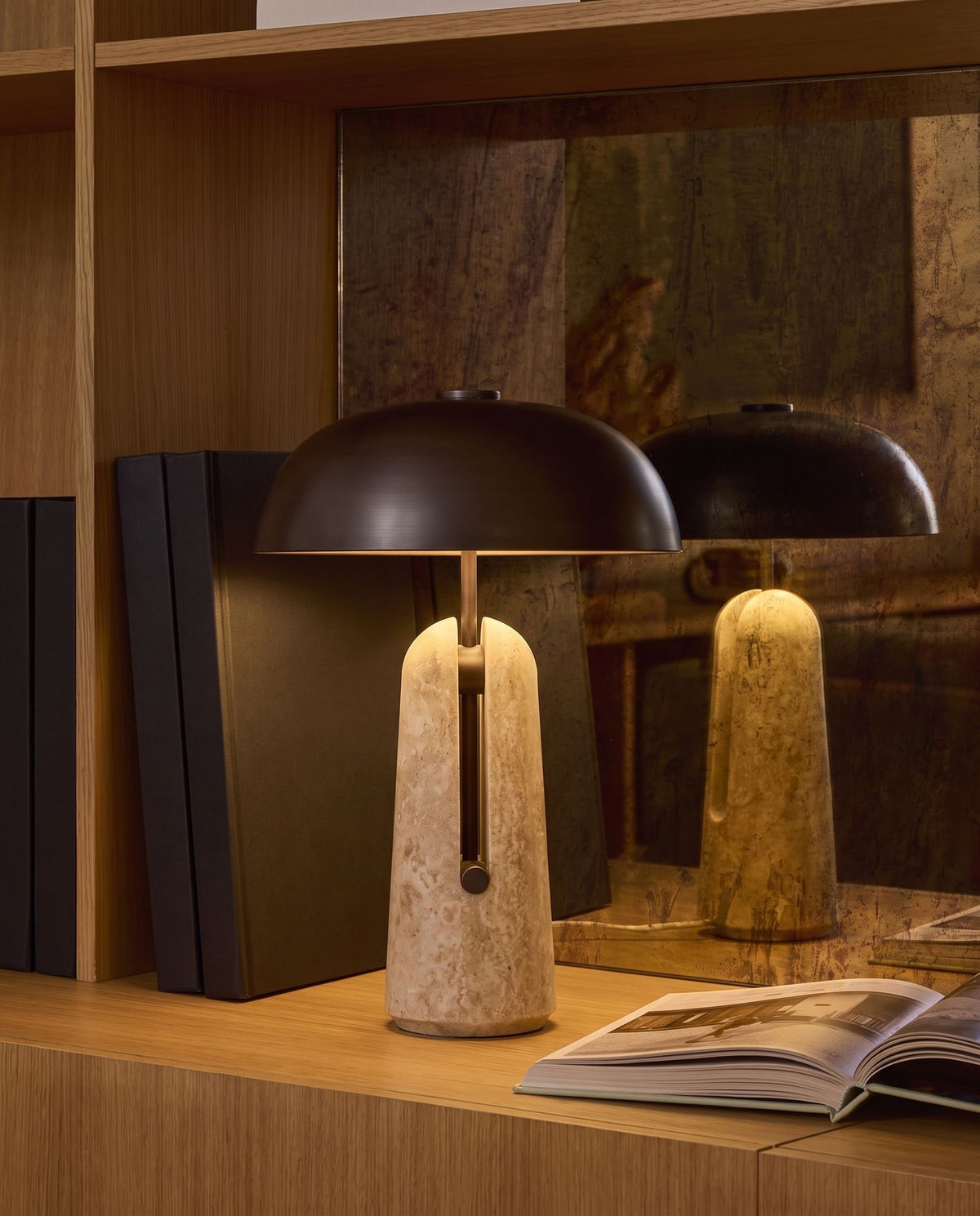 Sillon travertine and iron table lamp, lit, on an oak bookshelf before an antique mirror, open book