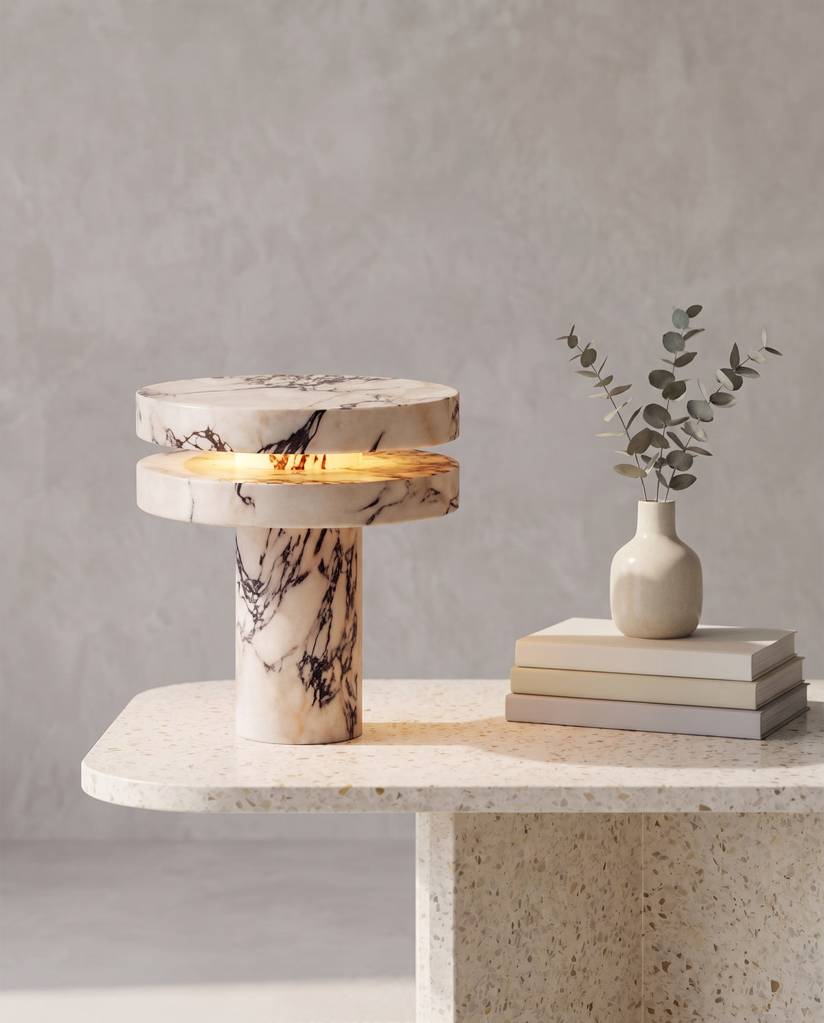 Strates marble table lamp, lit, on terrazzo table beside eucalyptus vase and books, grey plaster wall