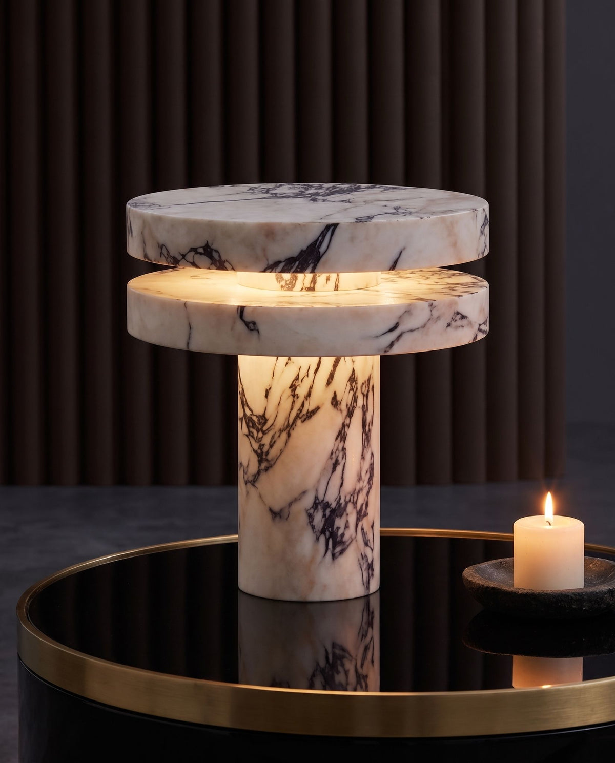 Strates marble table lamp, lit, on a black glass and brass coffee table, candle beside