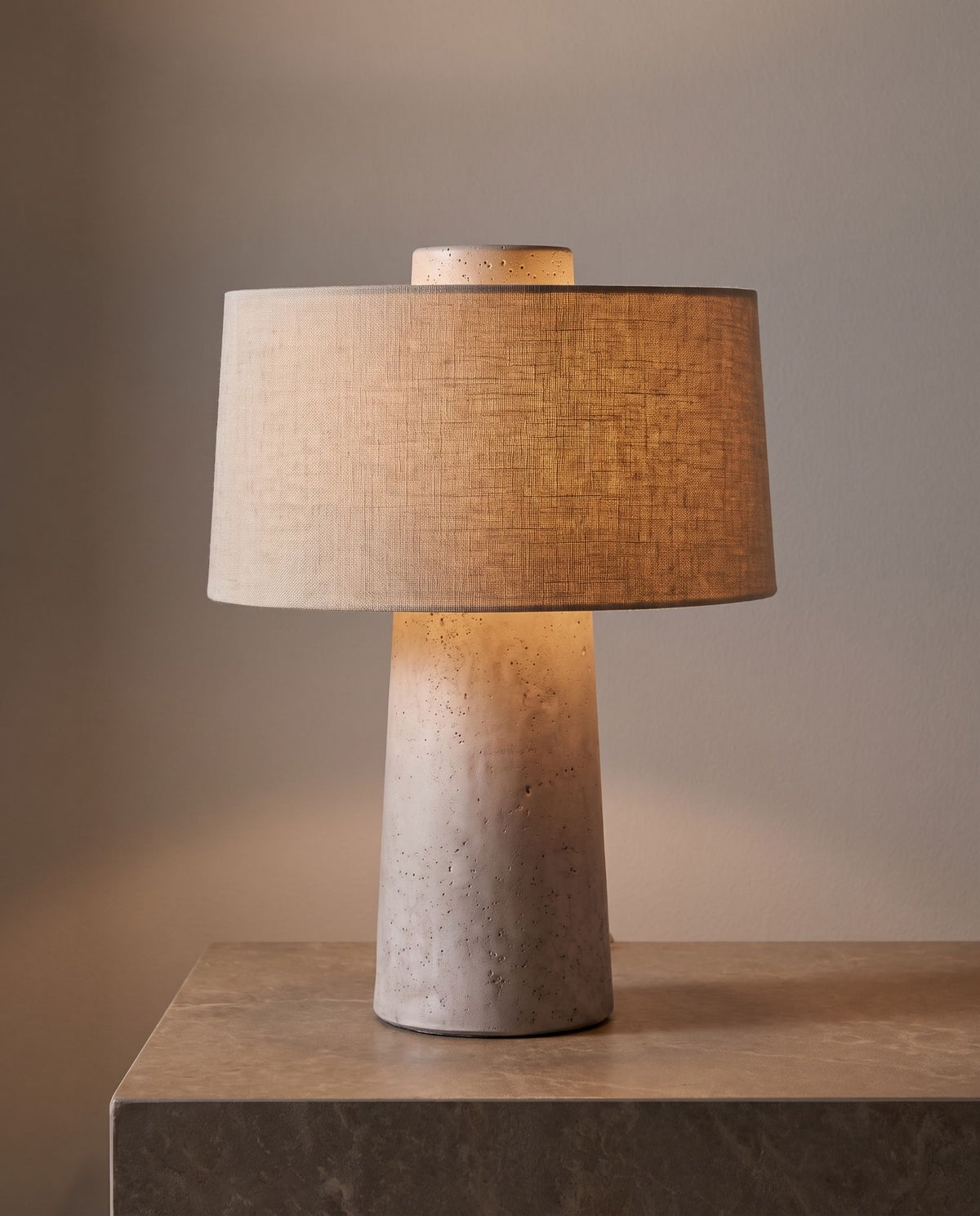 Tellur travertine and fabric table lamp, lit, against a soft taupe wall